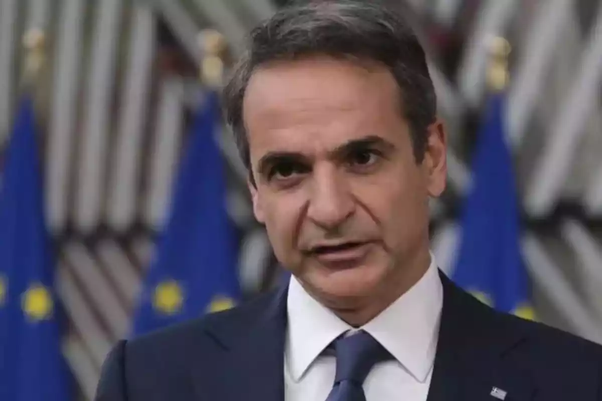 Hombre de cabello canoso con traje y corbata hablando frente a banderas de la Unión Europea desenfocadas al fondo