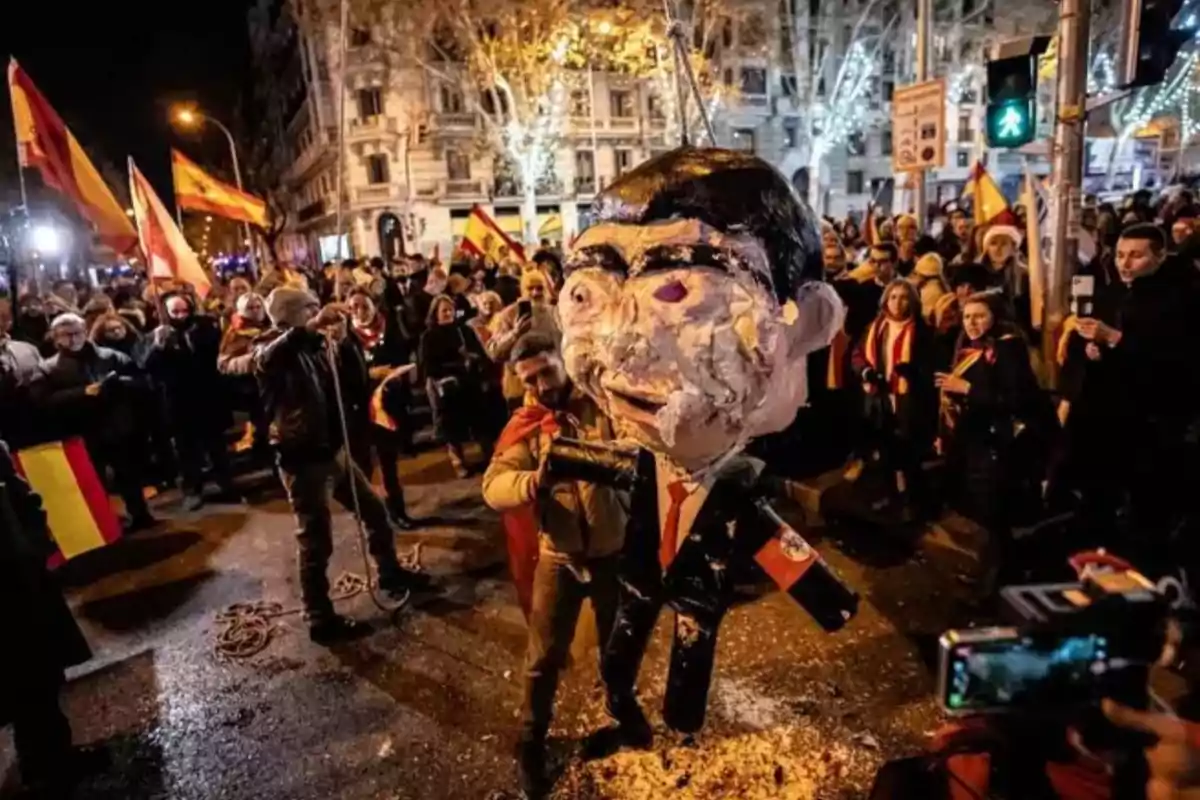 Manifestantes en una calle sostienen banderas de España y rodean una figura de cartón grande con el rostro cubierto de espuma o pintura durante una protesta nocturna