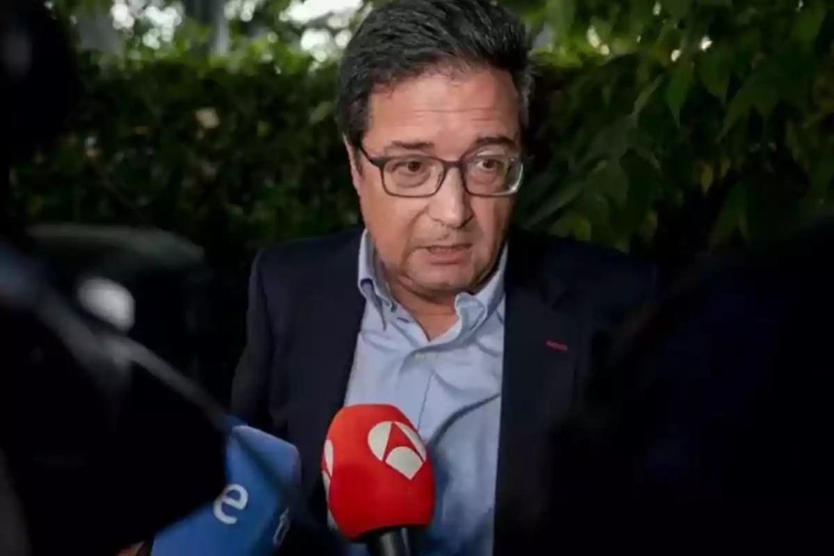 Hombre con gafas y saco oscuro hablando frente a varios micrófonos de prensa con fondo de vegetación