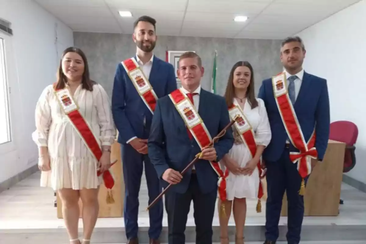 Cinco personas posan en un salón, todas llevan bandas rojas y blancas con un escudo, el hombre en el centro sostiene un bastón y todos visten ropa formal Cinco personas posan en un salón, todas llevan bandas rojas y blancas con un escudo, el hombre en el centro sostiene un bastón y todos visten ropa formal