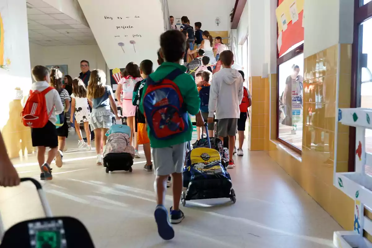 Niños entrando a la escuela con mochilas y maletas de ruedas en un pasillo iluminado Niños entrando a la escuela con mochilas y maletas de ruedas en un pasillo iluminado