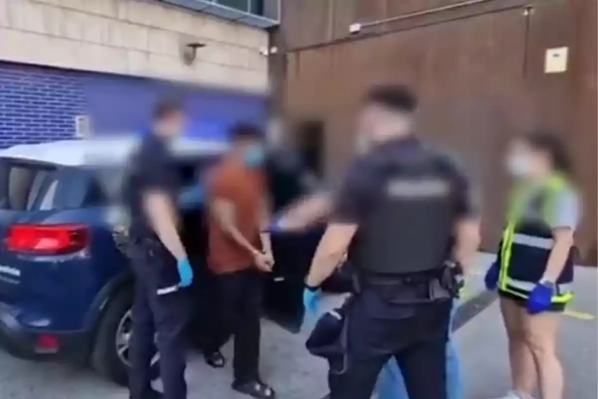 Varias personas con chalecos y guantes, aparentemente policías, rodean a un hombre esposado junto a un vehículo en la calle. Varias personas con chalecos y guantes, aparentemente policías, rodean a un hombre esposado junto a un vehículo en la calle.