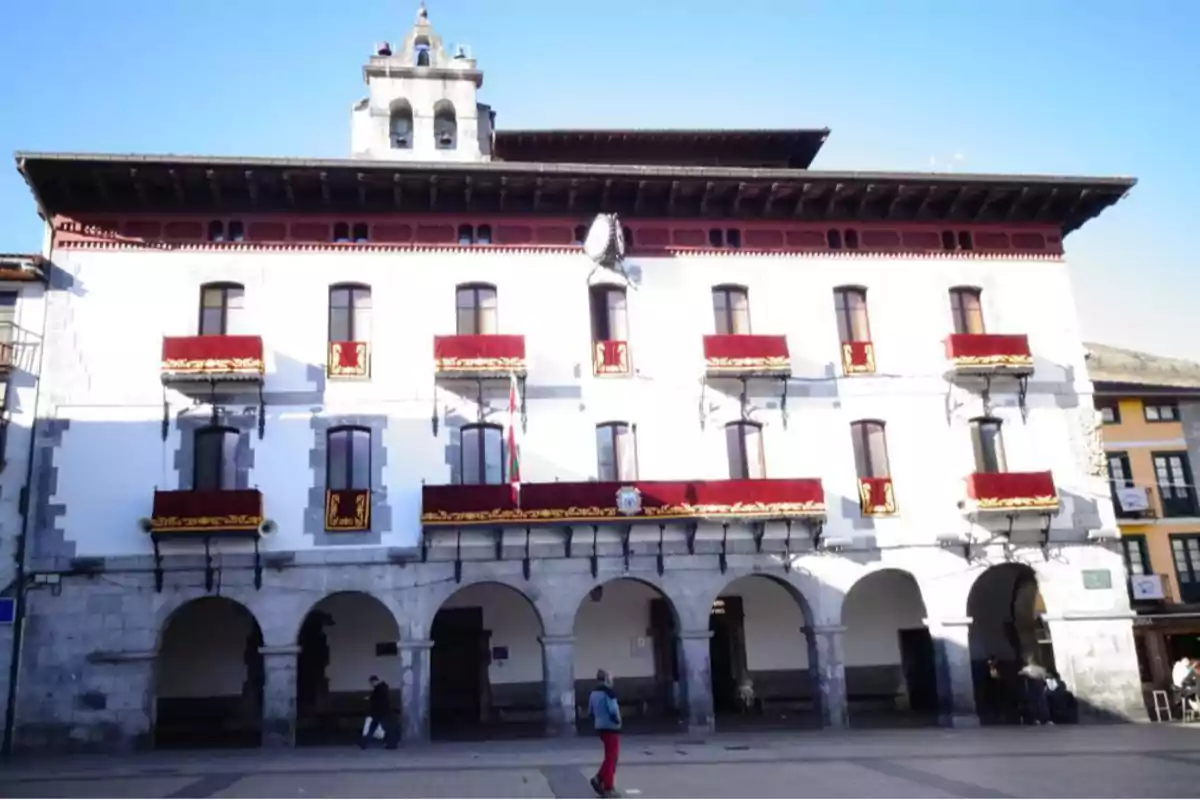 Fachada de un edificio histórico con balcones decorados con telas rojas y detalles dorados, arcos en la planta baja y una torre campanario en la parte superior Fachada de un edificio histórico con balcones decorados con telas rojas y detalles dorados, arcos en la planta baja y una torre campanario en la parte superior