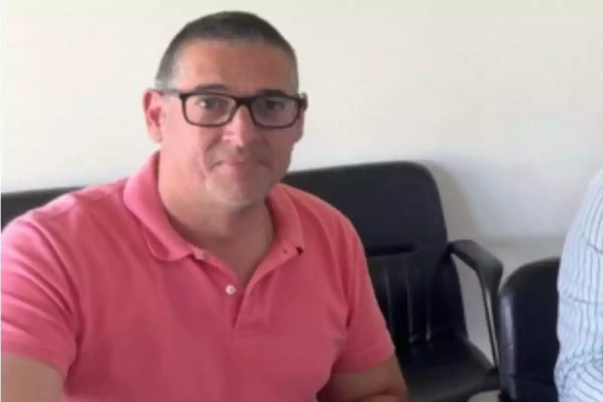 Hombre con gafas y camiseta rosa sentado en una sala con sillas negras al fondo Hombre con gafas y camiseta rosa sentado en una sala con sillas negras al fondo