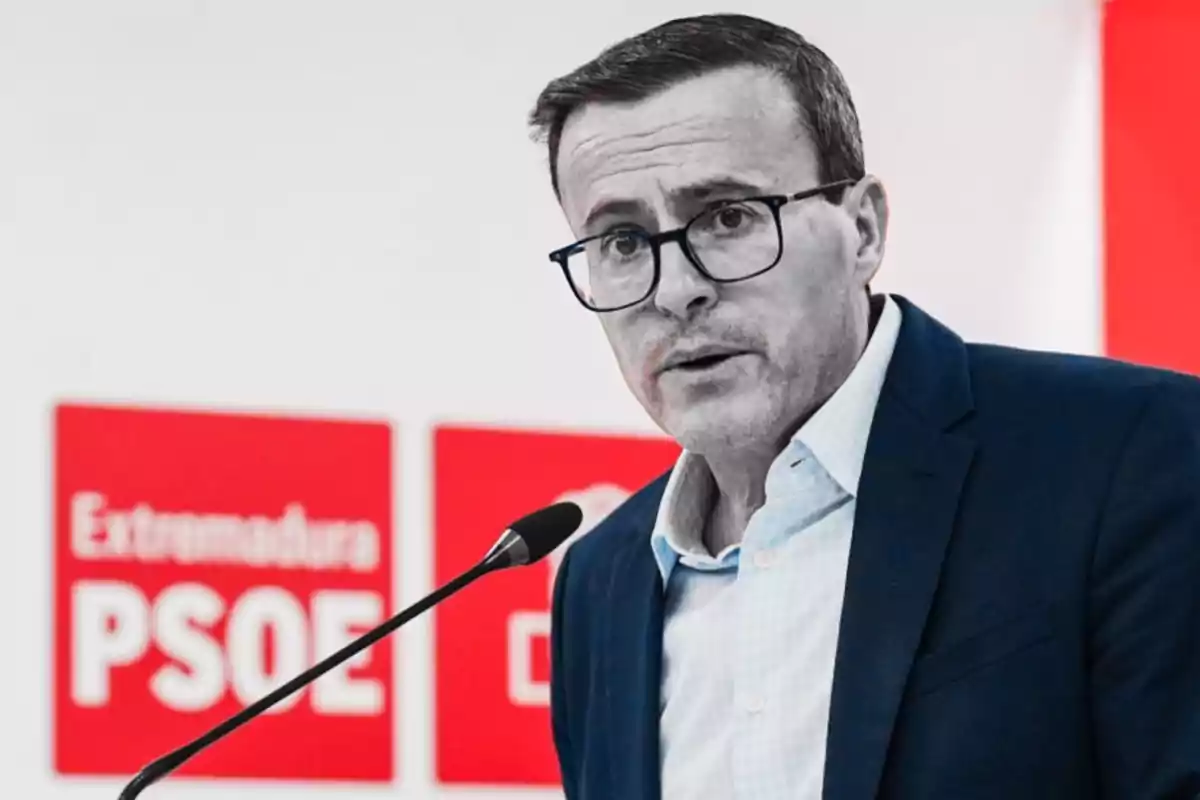 Hombre con gafas y traje azul hablando frente a un micrófono con carteles rojos de Extremadura PSOE al fondo