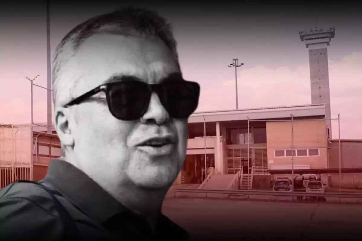 Hombre con gafas oscuras en primer plano frente a un edificio con torre de vigilancia y cercado
