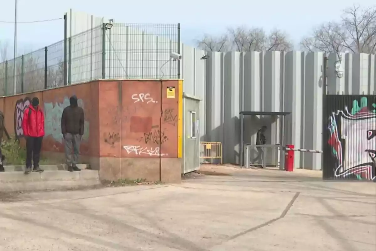 Personas de pie junto a un muro con grafitis y una valla metálica alta en un entorno urbano