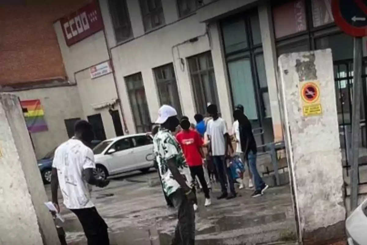 Un grupo de personas está reunido en la calle frente a un edificio con una bandera arcoíris colgada en la pared y varios carteles visibles Un grupo de personas está reunido en la calle frente a un edificio con una bandera arcoíris colgada en la pared y varios carteles visibles