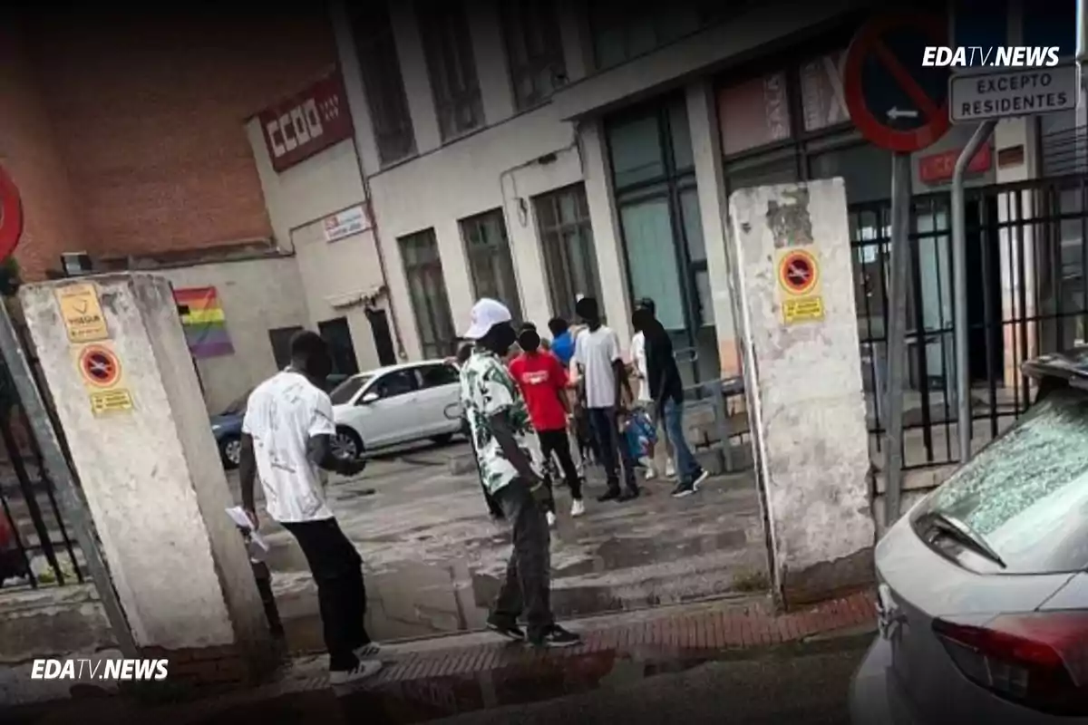 Un grupo de personas camina y conversa cerca de la entrada de un edificio en una calle urbana con varios carteles y una bandera arcoíris en la pared de fondo