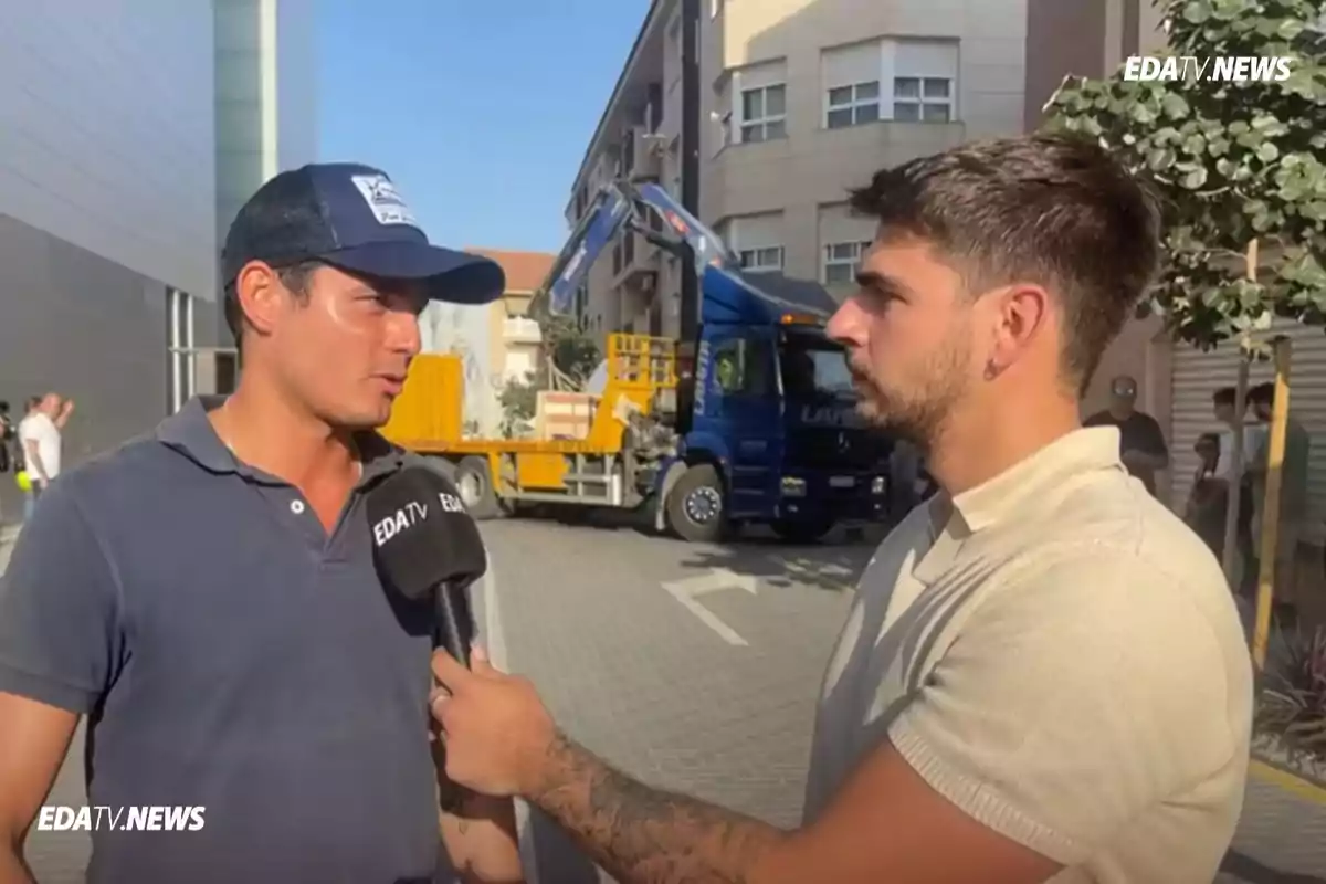 Un reportero entrevista a un hombre con gorra en la calle mientras un camión azul y amarillo se encuentra estacionado al fondo