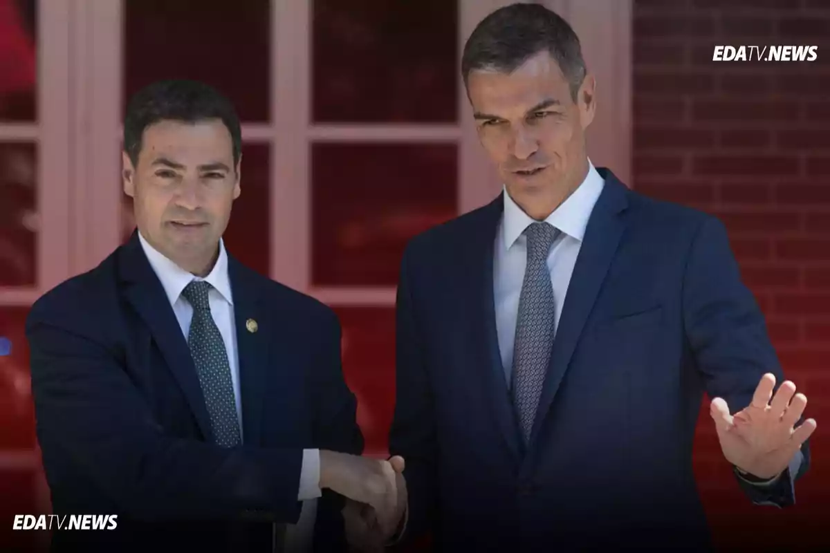 Dos hombres con traje se dan la mano frente a una ventana mientras uno de ellos saluda con la otra mano