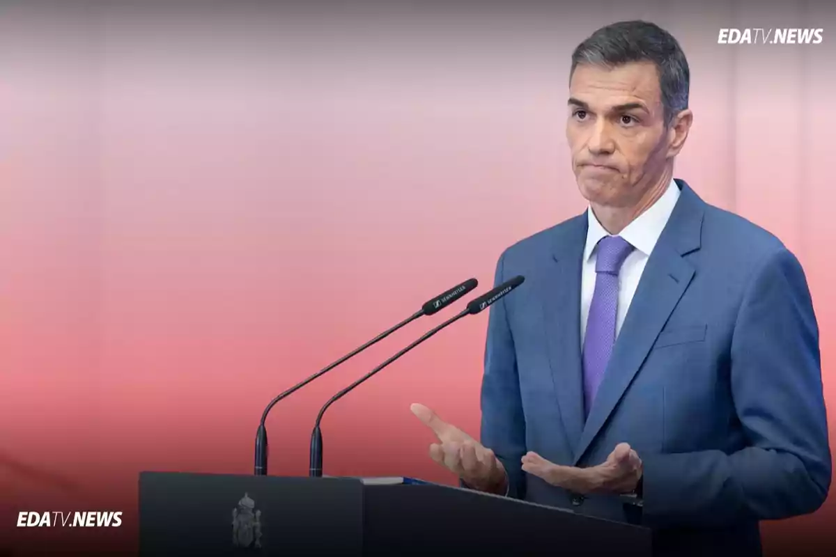 Hombre con traje azul y corbata morada hablando en una conferencia de prensa con gesto de incertidumbre frente a dos micrófonos y fondo rosado