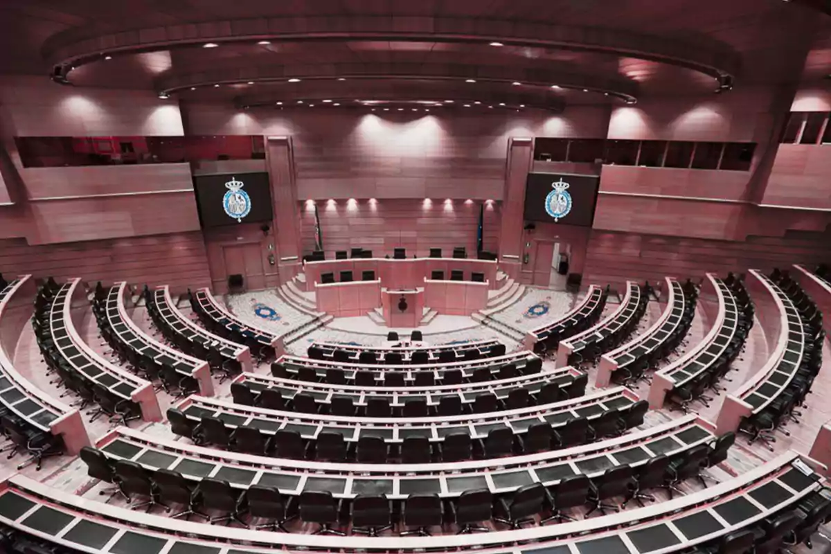 Vista panorámica de un moderno hemiciclo parlamentario con asientos vacíos y dos pantallas grandes mostrando un escudo oficial