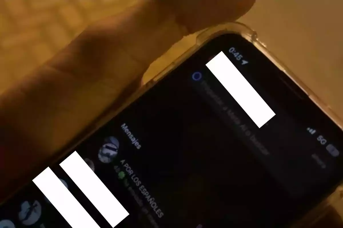 Persona sosteniendo un teléfono móvil con la pantalla encendida mostrando una aplicación de mensajería en modo oscuro y algunos textos y nombres cubiertos por recuadros blancos Persona sosteniendo un teléfono móvil con la pantalla encendida mostrando una aplicación de mensajería en modo oscuro y algunos textos y nombres cubiertos por recuadros blancos