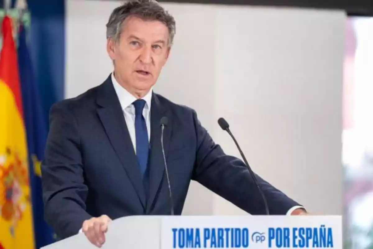 Hombre de traje azul hablando en un atril con el texto Toma partido por España y banderas al fondo