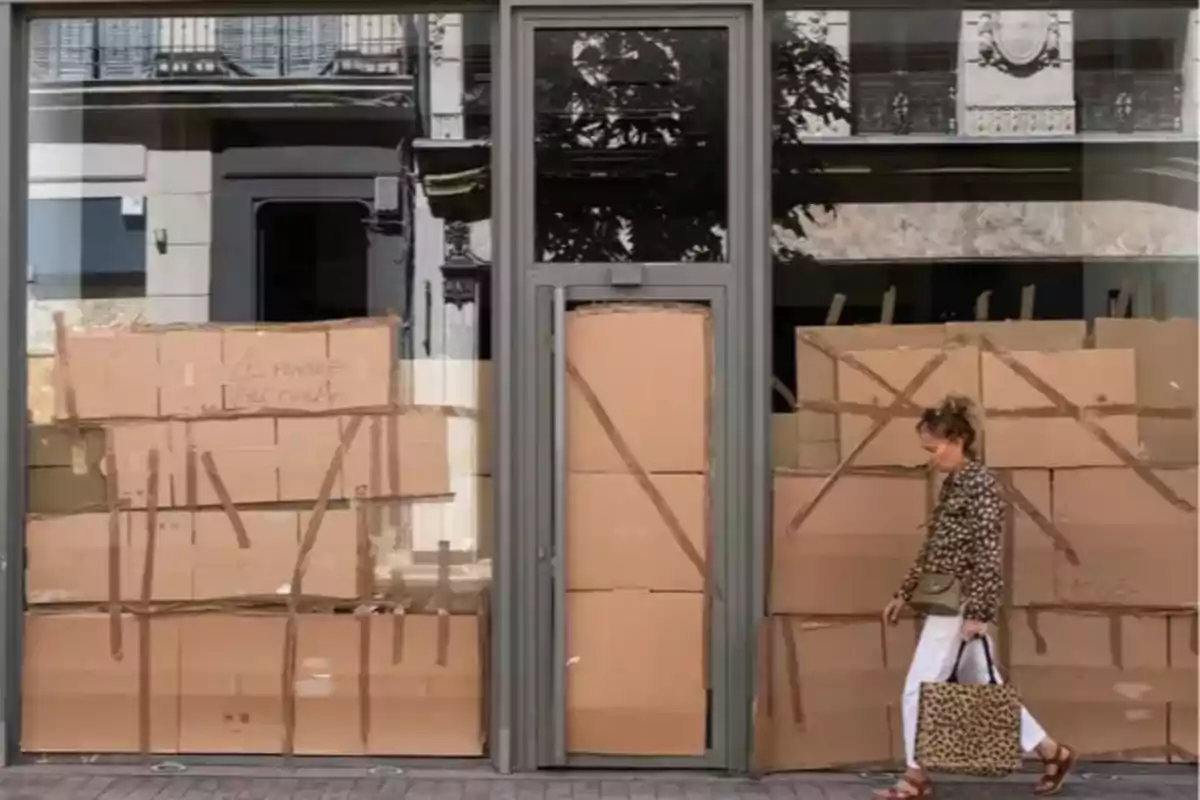Mujer caminando frente a una tienda con ventanas y puerta cubiertas con cajas de cartón sujetas con cinta adhesiva Mujer caminando frente a una tienda con ventanas y puerta cubiertas con cajas de cartón sujetas con cinta adhesiva