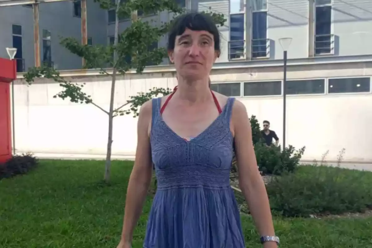 Mujer de cabello corto y oscuro con vestido azul de tirantes posando en un jardín frente a un edificio moderno