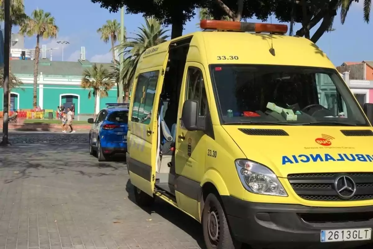 Ambulancia amarilla estacionada con la puerta lateral abierta en una calle junto a un coche de policía y palmeras al fondo Ambulancia amarilla estacionada con la puerta lateral abierta en una calle junto a un coche de policía y palmeras al fondo