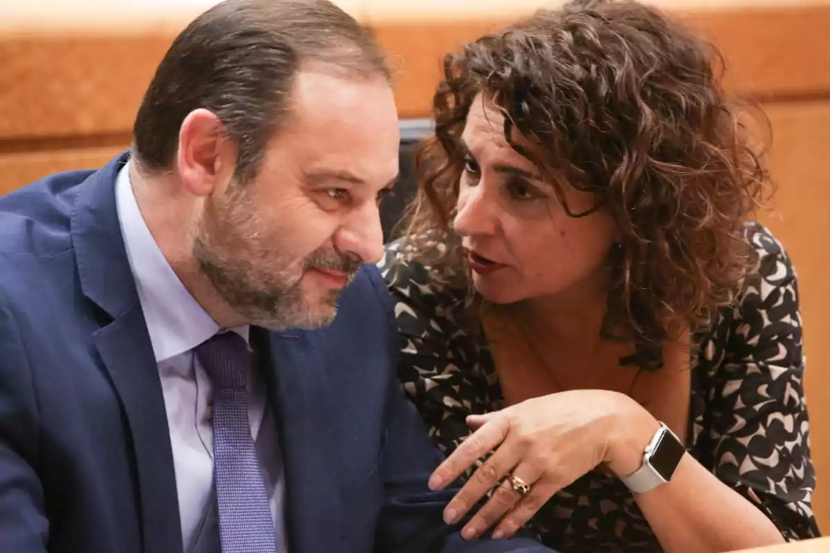 Dos personas conversan de cerca en un entorno formal, una de ellas lleva traje y la otra un vestido estampado y reloj inteligente. Dos personas conversan de cerca en un entorno formal, una de ellas lleva traje y la otra un vestido estampado y reloj inteligente.
