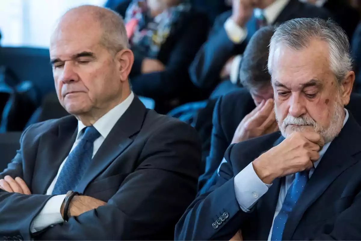 Dos hombres mayores con expresión seria y pensativa, vestidos con traje y corbata, sentados en lo que parece ser una sala de reuniones o juicio.