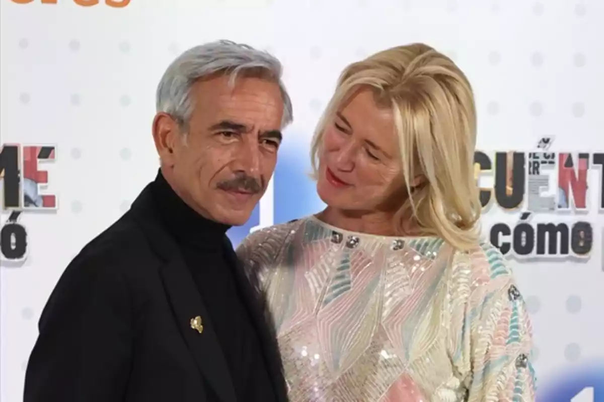 Una pareja posa sonriente en un evento con fondo de letras y colores claros Una pareja posa sonriente en un evento con fondo de letras y colores claros