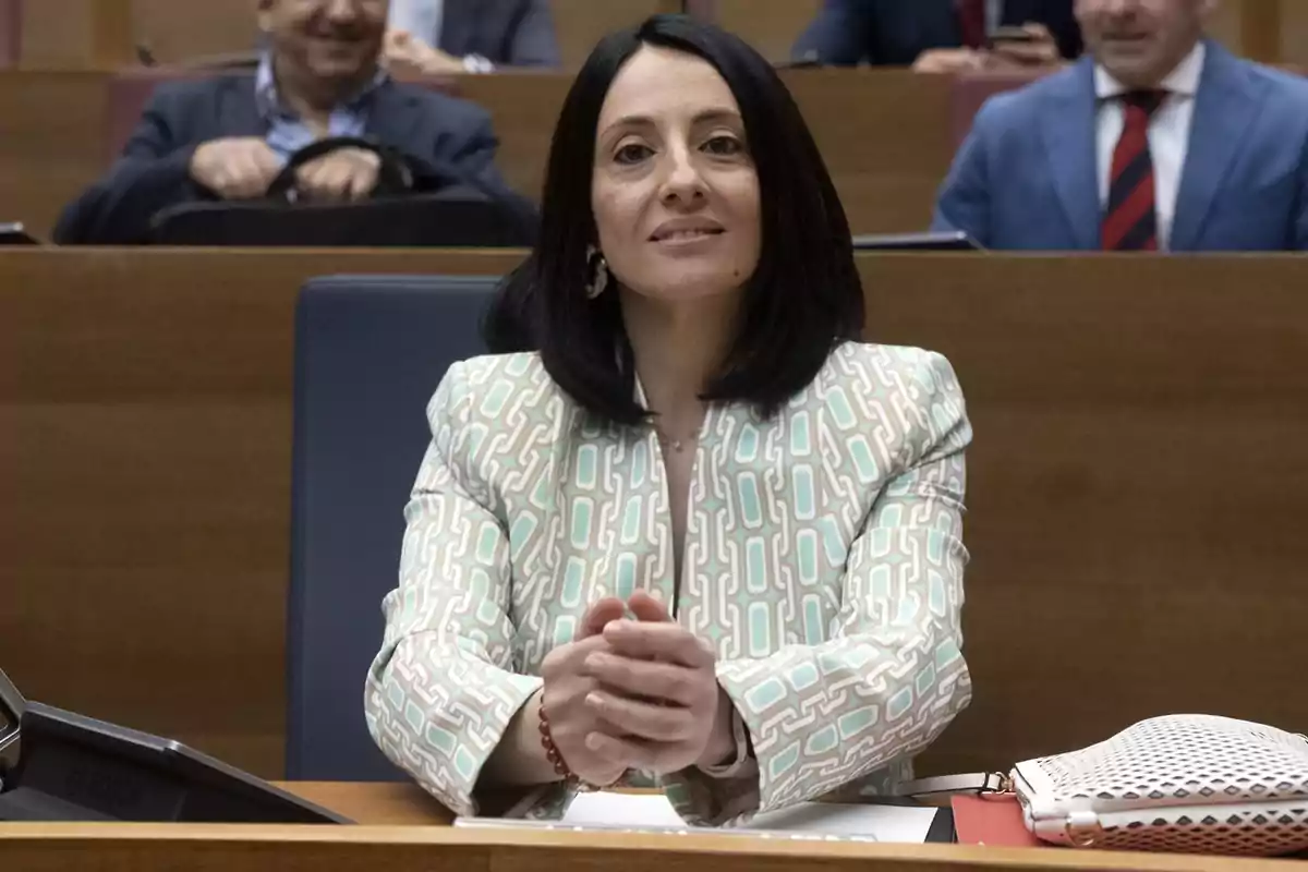 Mujer de cabello oscuro sentada en una sala de reuniones con las manos entrelazadas sobre la mesa y una chaqueta de colores claros con estampado geométrico Mujer de cabello oscuro sentada en una sala de reuniones con las manos entrelazadas sobre la mesa y una chaqueta de colores claros con estampado geométrico