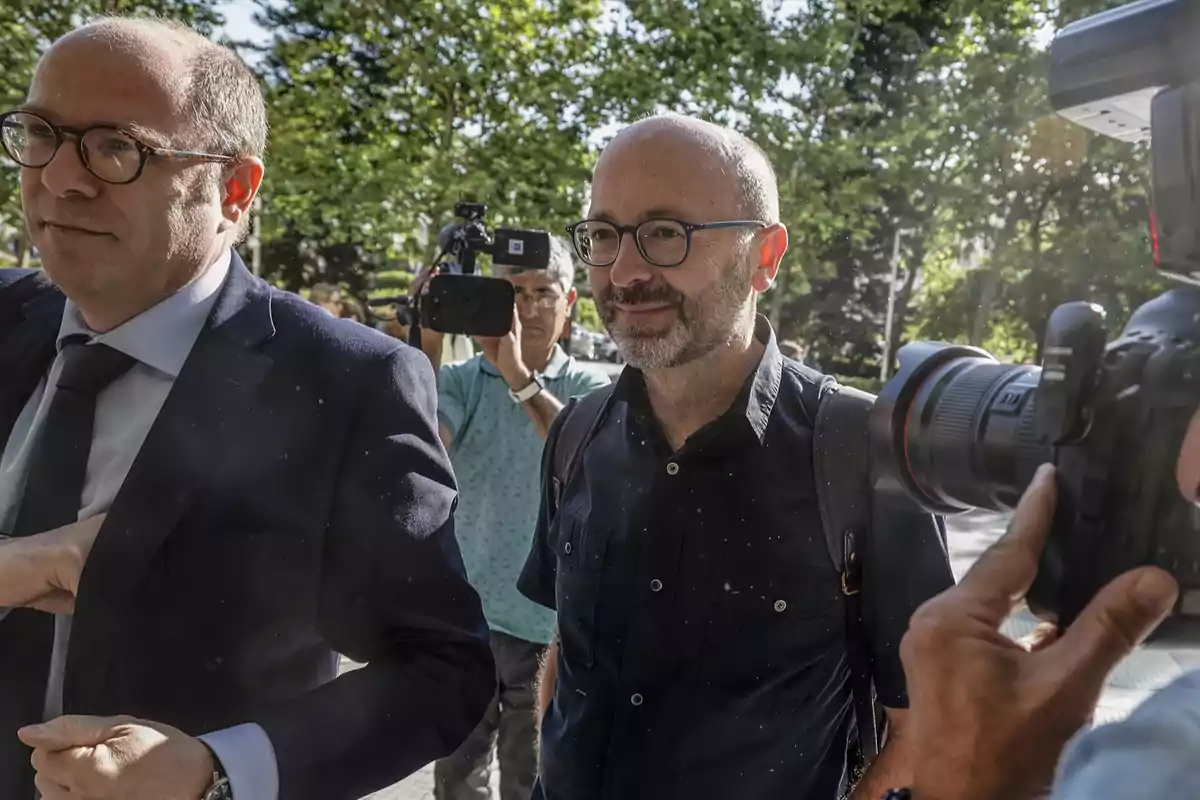 Dos hombres caminando al aire libre mientras son fotografiados y grabados por periodistas con cámaras profesionales en un entorno urbano con árboles al fondo