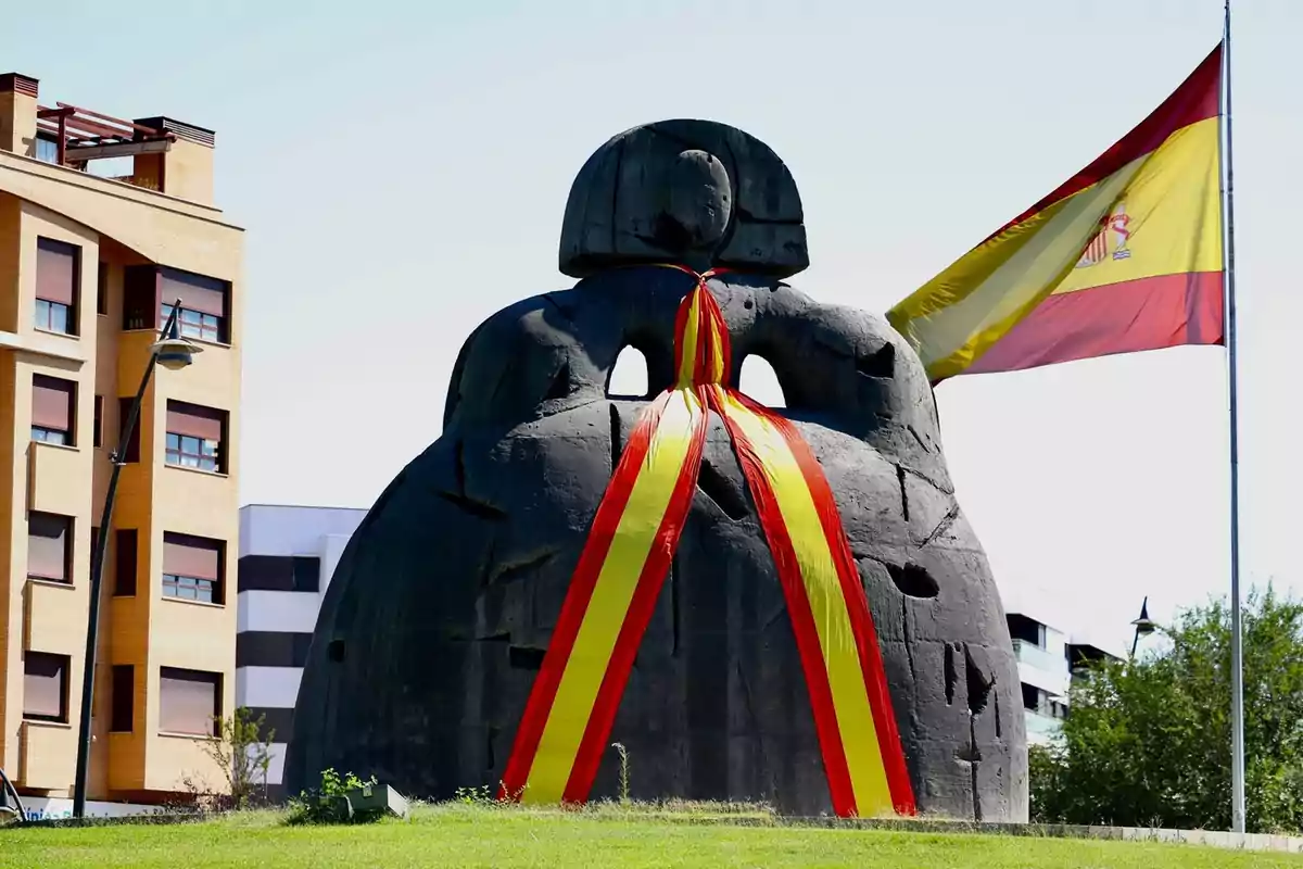 Escultura negra de gran tamaño decorada con una cinta roja y amarilla junto a una bandera de España ondeando y edificios residenciales al fondo