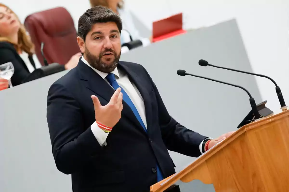 Hombre con barba y traje azul hablando en un podio con micrófonos en un entorno institucional mientras personas escuchan al fondo Hombre con barba y traje azul hablando en un podio con micrófonos en un entorno institucional mientras personas escuchan al fondo