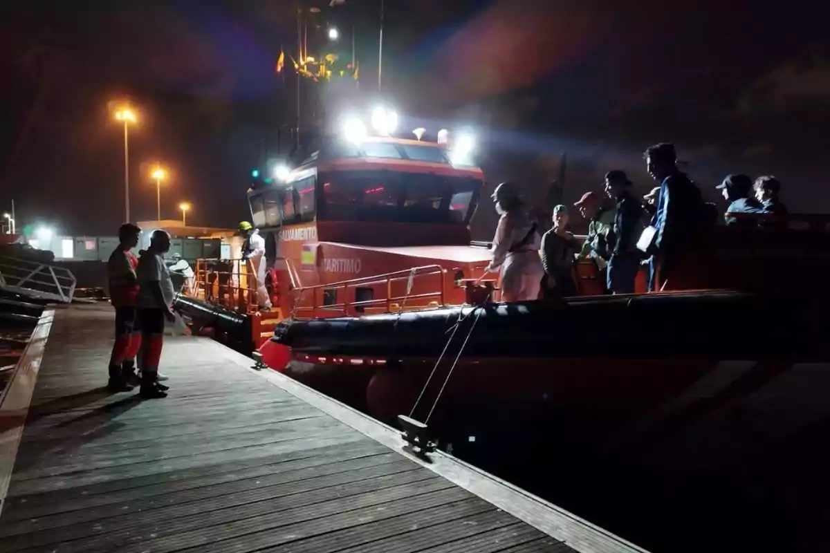 Un grupo de personas desembarca de una embarcación de salvamento marítimo iluminada en un muelle durante la noche Un grupo de personas desembarca de una embarcación de salvamento marítimo iluminada en un muelle durante la noche