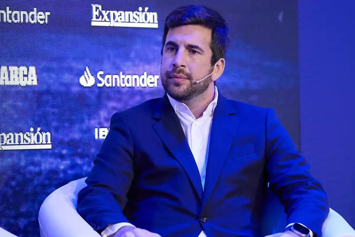 Hombre con barba y cabello corto usando un traje azul y camisa blanca sentado en un sillón blanco durante un evento con fondo de logotipos de empresas. Hombre con barba y cabello corto usando un traje azul y camisa blanca sentado en un sillón blanco durante un evento con fondo de logotipos de empresas.