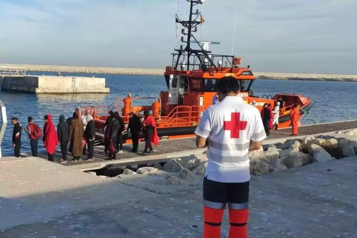 Un grupo de personas desembarca de un barco de rescate naranja en un puerto mientras un miembro de la Cruz Roja observa desde el muelle.