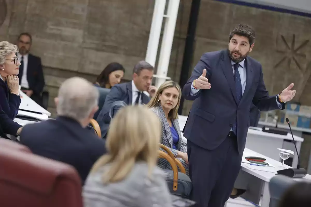 Un hombre de traje habla gesticulando con las manos en una reunión formal mientras varias personas lo escuchan atentamente en una sala de conferencias