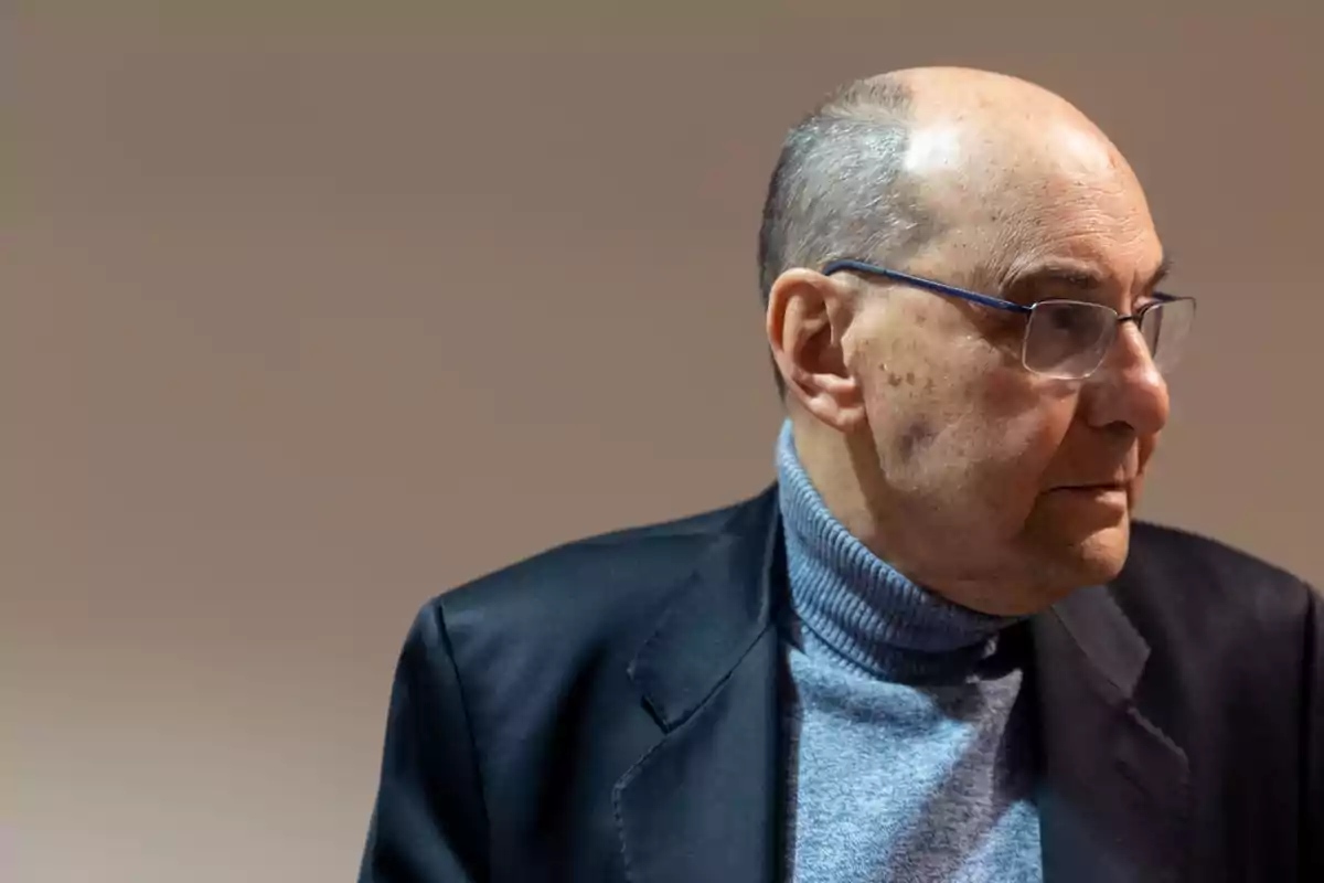 Hombre mayor con gafas y suéter gris mirando hacia la derecha con fondo liso beige