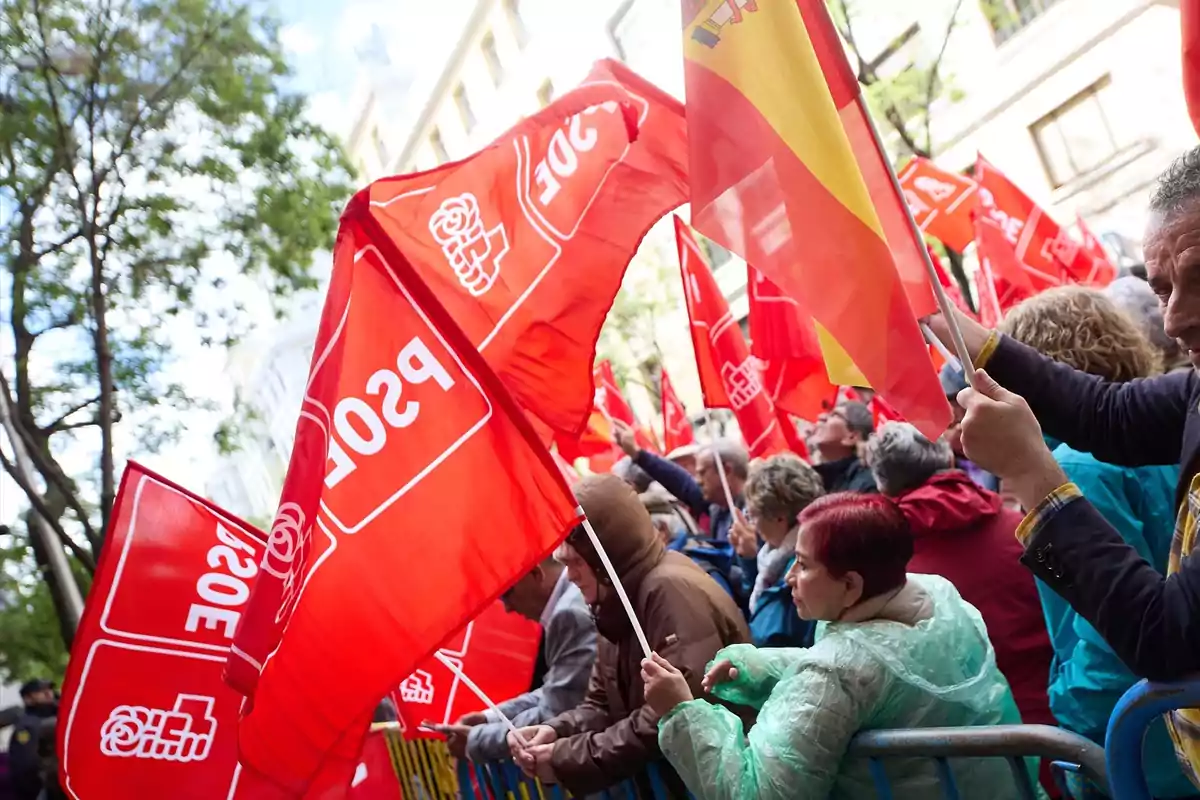 Personas en una manifestación sosteniendo banderas rojas del PSOE y una bandera de España en una calle rodeada de edificios y árboles