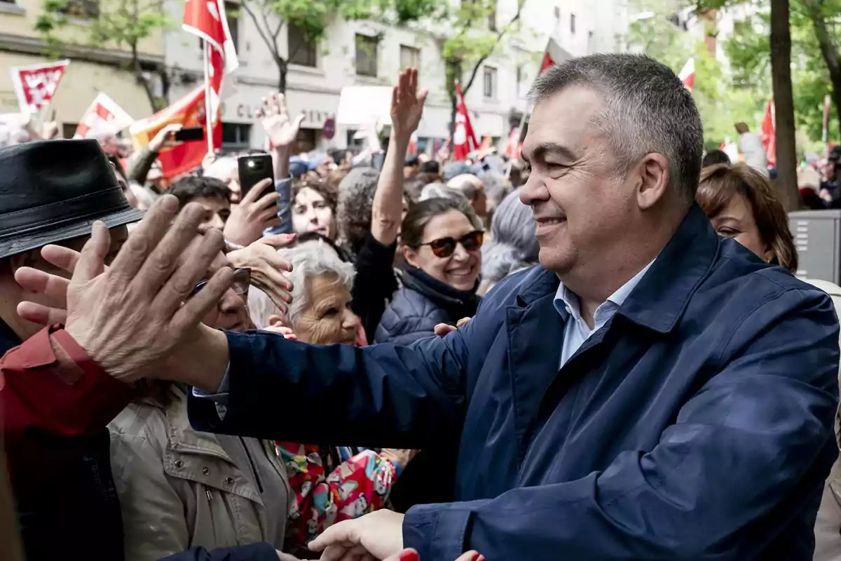 Hombre sonriente con chaqueta azul saluda a varias personas en una multitud durante un evento al aire libre con banderas y ambiente festivo Hombre sonriente con chaqueta azul saluda a varias personas en una multitud durante un evento al aire libre con banderas y ambiente festivo