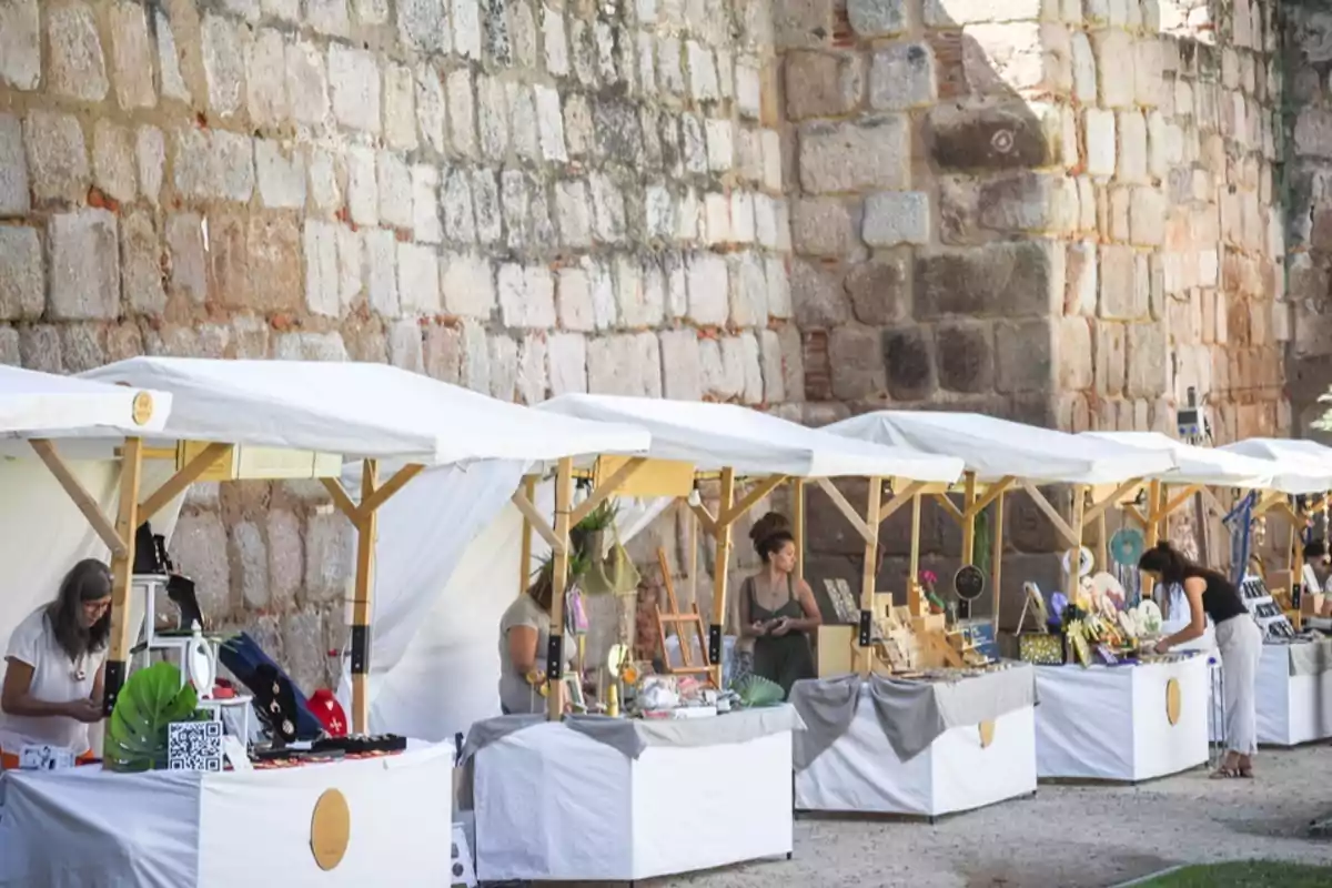 Puestos de mercado artesanal al aire libre con varias personas organizando productos bajo toldos blancos frente a una pared de piedra Puestos de mercado artesanal al aire libre con varias personas organizando productos bajo toldos blancos frente a una pared de piedra