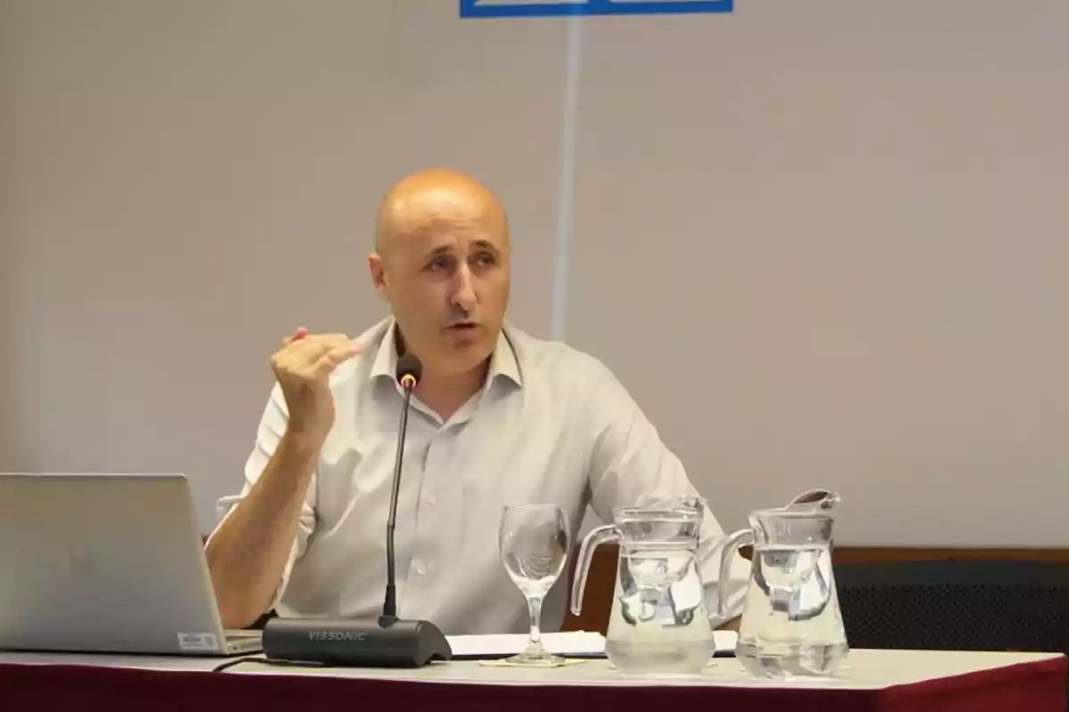 Hombre calvo con camisa clara hablando en una conferencia sentado frente a una mesa con una laptop, una copa y dos jarras de agua