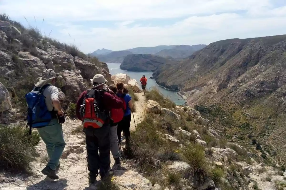 Grupo de personas haciendo senderismo por un sendero estrecho en la montaña con vista a un río y montañas al fondo
