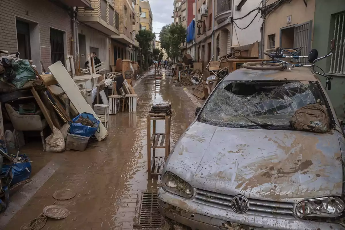 Calle llena de lodo y escombros con un coche dañado y muebles apilados tras una inundación