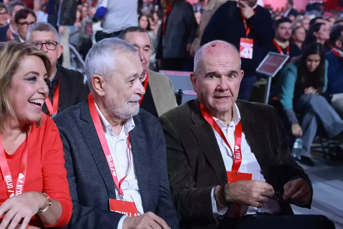 Tres personas sentadas en primera fila en un evento con credenciales rojas rodeadas de más asistentes en un ambiente animado Tres personas sentadas en primera fila en un evento con credenciales rojas rodeadas de más asistentes en un ambiente animado