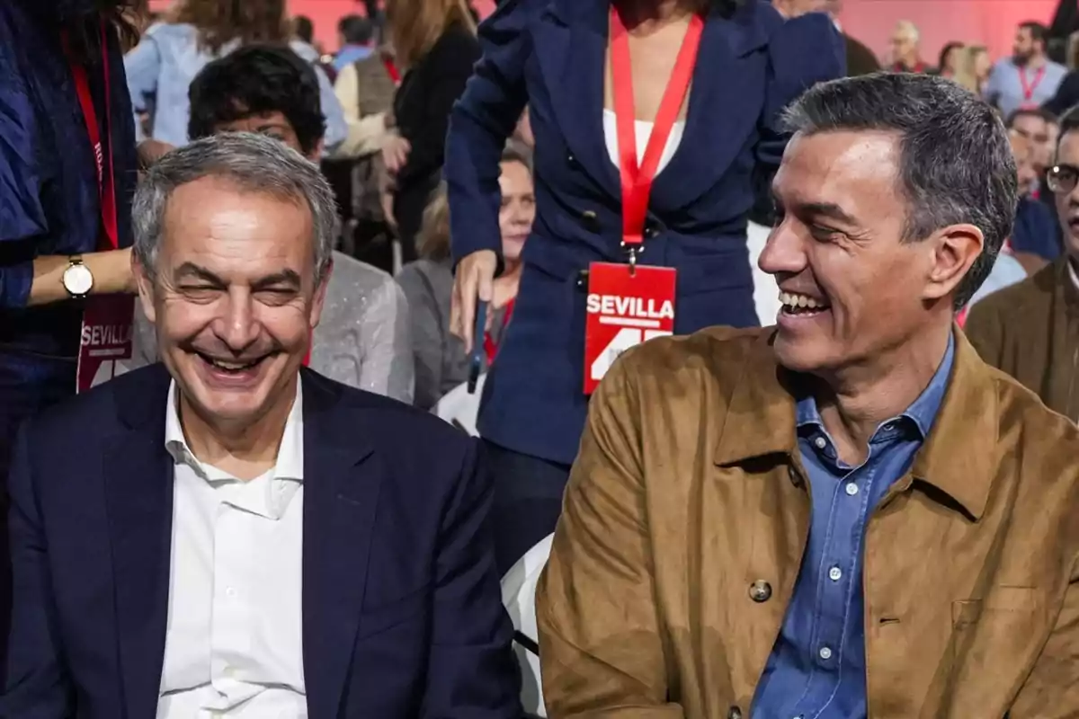 Dos hombres sonríen y conversan sentados en un evento con varias personas al fondo y una mujer de pie con una acreditación roja que dice Sevilla.