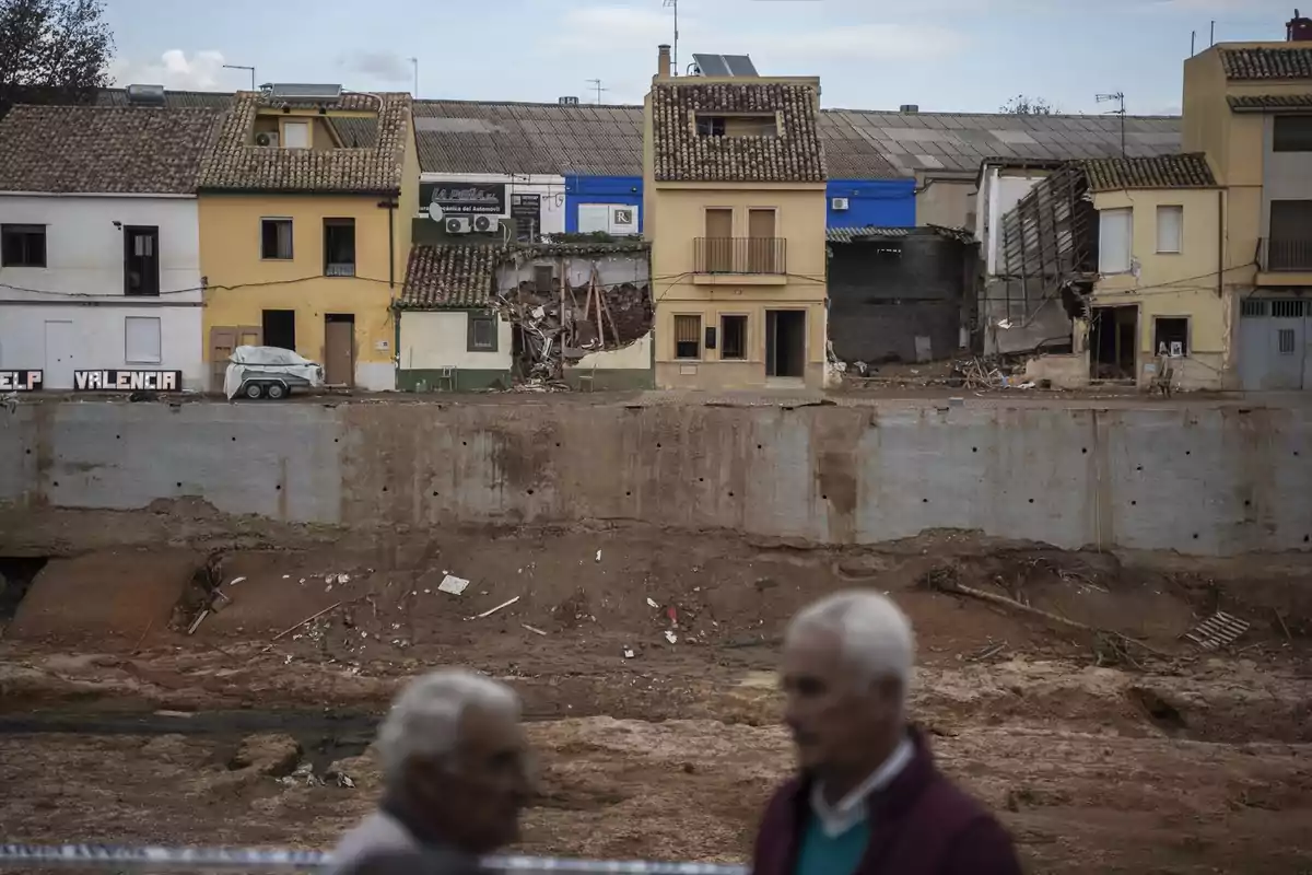 Casas dañadas y parcialmente derrumbadas junto a una zanja profunda con dos personas mayores desenfocadas en primer plano Casas dañadas y parcialmente derrumbadas junto a una zanja profunda con dos personas mayores desenfocadas en primer plano
