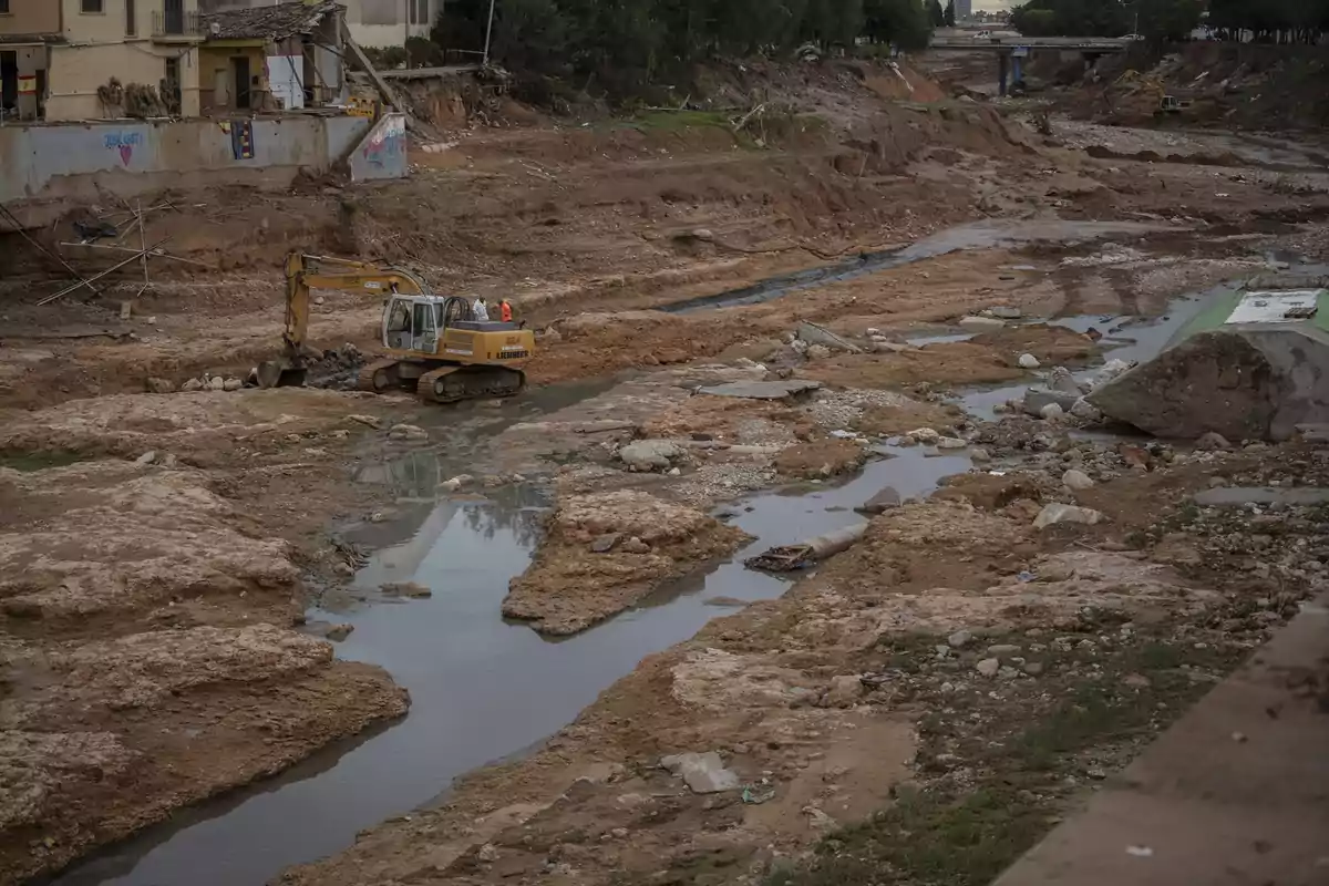 Excavadora trabajando en un cauce seco y erosionado con charcos de agua y escombros, cerca de una zona urbana dañada Excavadora trabajando en un cauce seco y erosionado con charcos de agua y escombros, cerca de una zona urbana dañada