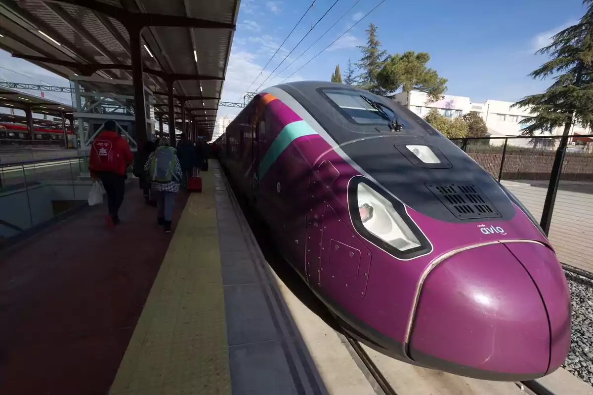 Tren de alta velocidad color morado estacionado en un andén con personas caminando junto a él Tren de alta velocidad color morado estacionado en un andén con personas caminando junto a él