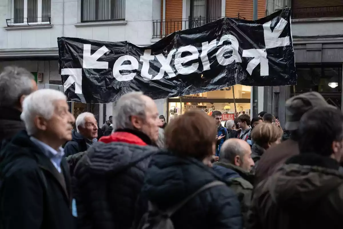 Varias personas reunidas en una manifestación en la calle con una pancarta negra que tiene la palabra etxera escrita en blanco y flechas apuntando hacia el centro