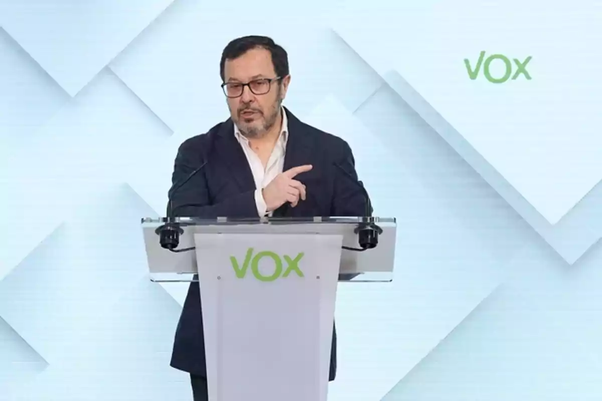 Hombre con gafas y barba hablando en un atril con el logo de VOX sobre un fondo claro geométrico Hombre con gafas y barba hablando en un atril con el logo de VOX sobre un fondo claro geométrico