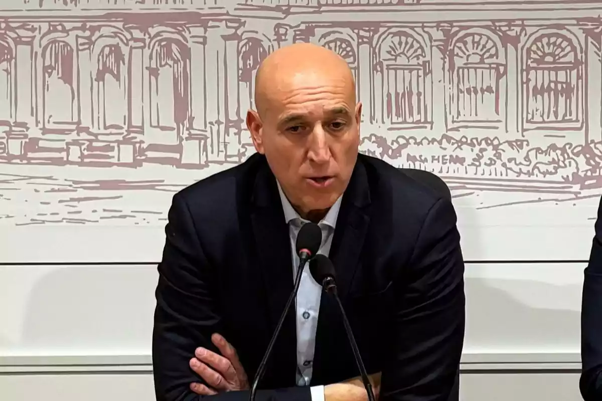 Hombre calvo con traje oscuro hablando frente a dos micrófonos en una conferencia de prensa con fondo de ilustración arquitectónica