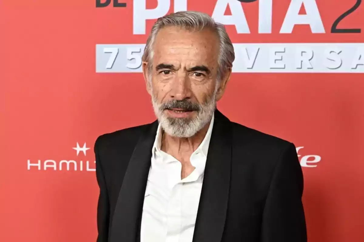 Hombre de cabello y barba canosos con saco negro y camisa blanca posando frente a un fondo rojo con letras blancas
