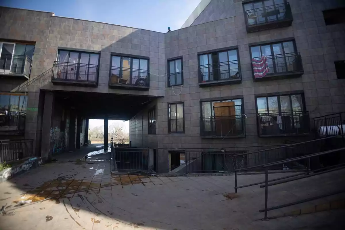 Edificio de apartamentos de fachada gris con balcones y ventanas, algunas con ropa colgada, y una rampa en primer plano Edificio de apartamentos de fachada gris con balcones y ventanas, algunas con ropa colgada, y una rampa en primer plano